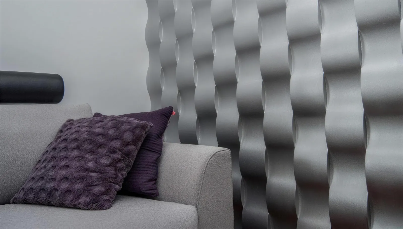 Szara sofa z fioletową poduszką przy ścianie z przestrzennych srebrnych paneli